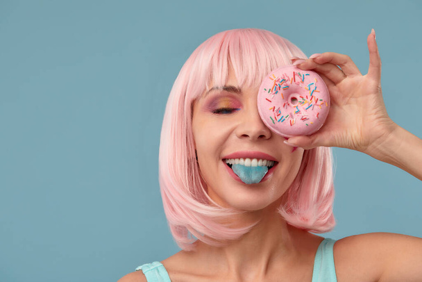 Portret van een modieus meisje in een roze pruik en met een roze donut in haar handen houdt het oog, toont tong - Foto, afbeelding