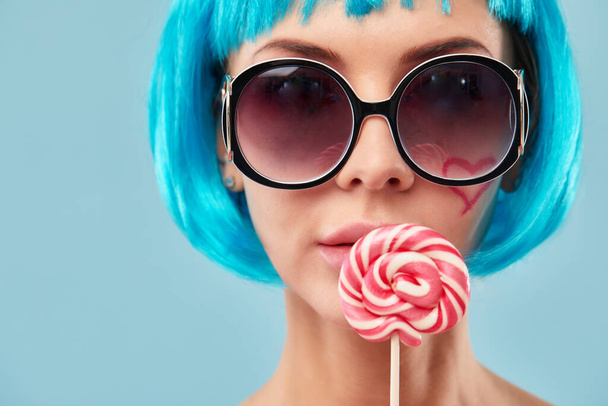 Young funky girl in blue wig and eyeglasses, holding lollipop candy, looking aside with dreamful smile as if making plans for party, isolated on blue background - Фото, изображение