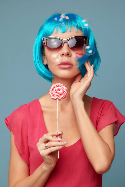 Young funky girl in blue wig and eyeglasses, holding lollipop candy, looking aside with dreamful smile as if making plans for party, isolated on blue background - Fotó, kép
