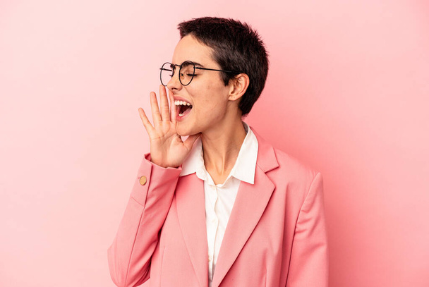 Joven mujer de negocios con una chaqueta rosa aislada sobre fondo rosa gritando y sosteniendo la palma cerca de la boca abierta. - Foto, imagen