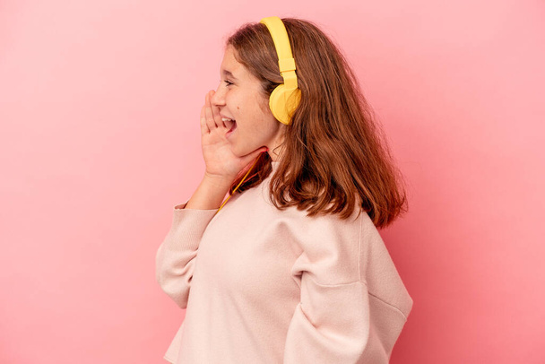 Little caucasian girl listening to music isolated on pink background shouting and holding palm near opened mouth. - Zdjęcie, obraz