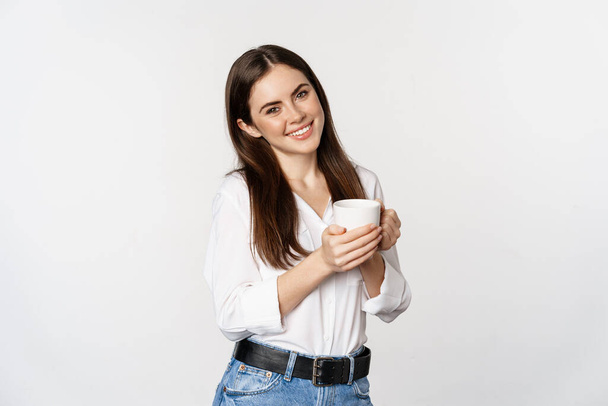 Corporate woman standing with coffee tea mug and smiling, drinking from cup, standing happy against white background - Foto, imagen