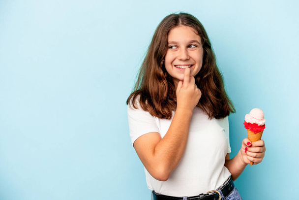 Kleines kaukasisches Mädchen isst ein Eis isoliert auf blauem Hintergrund und denkt entspannt über etwas nach, das einen Kopierraum betrachtet. - Foto, Bild