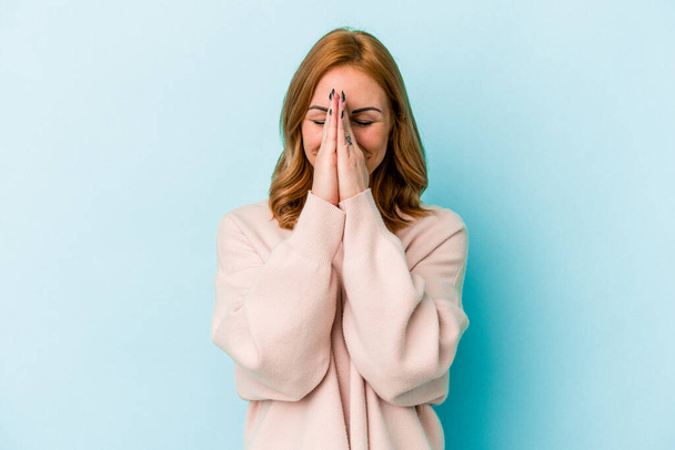 Young caucasian woman isolated on blue background holding hands in pray near mouth, feels confident. - Foto, afbeelding