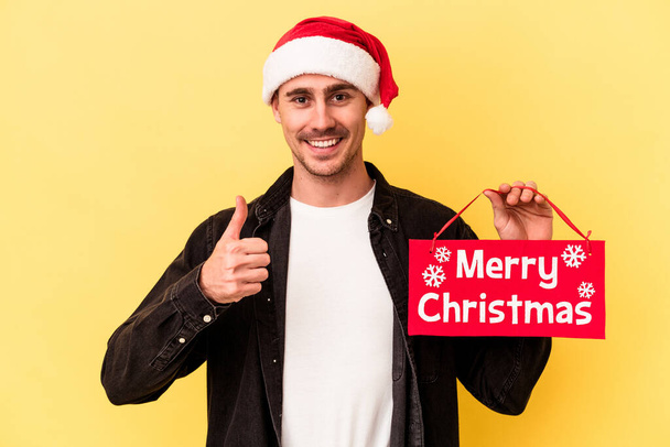 Młody biały mężczyzna z tabliczką Wesołych Świąt odizolowany na żółtym tle uśmiecha się i podnosi kciuk w górę - Zdjęcie, obraz