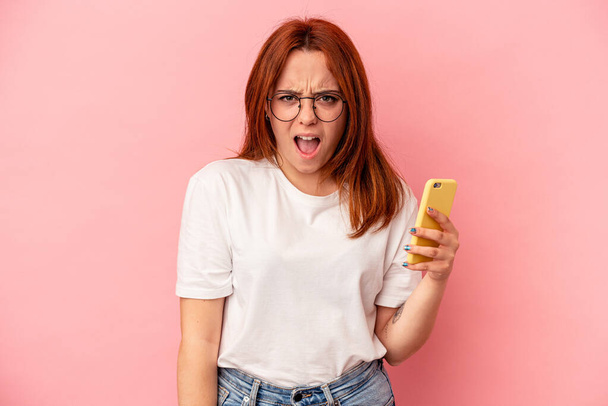Mladá běloška drží mobilní telefon izolované na růžovém pozadí křičí velmi naštvaný a agresivní. - Fotografie, Obrázek