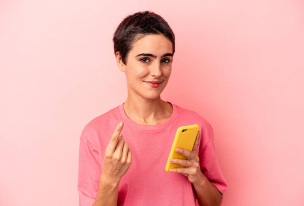 Νεαρή καυκάσια γυναίκα κρατώντας το κινητό τηλέφωνο απομονωμένο σε ροζ φόντο δείχνοντας με το δάχτυλο σε σας σαν να προσκαλώντας έρθει πιο κοντά. - Φωτογραφία, εικόνα