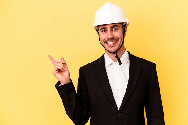 Young architect caucasian man isolated on yellow background smiling and pointing aside, showing something at blank space. - Photo, Image