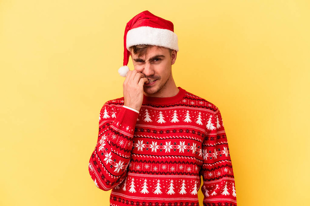 若いです白人男性祝うクリスマスは黄色の背景に隔離された爪をかむ,神経と非常に不安. - 写真・画像