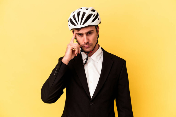 Nuori valkoihoinen liikemies pyöräilykypärä päässä eristettynä keltaisella pohjalla osoittaen sormella ohimoa, keskittyen tehtävään. - Valokuva, kuva