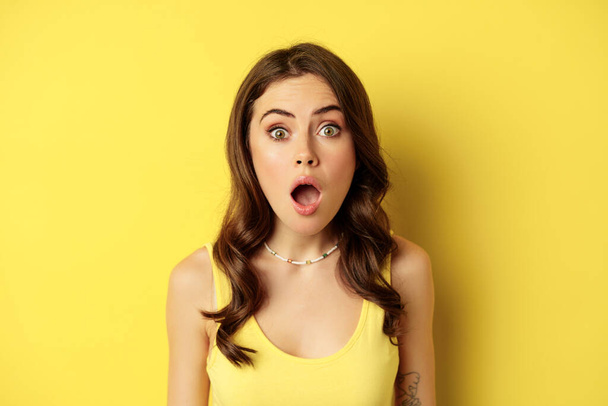 Close up portrait of stylish brunette girl looking amazed, wow face, showing excitement and surprise, impressed by something, standing over yellow background - Fotoğraf, Görsel
