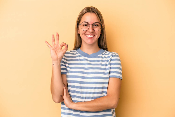Young caucasian woman isolated on yellow background winks an eye and holds an okay gesture with hand. - Photo, Image