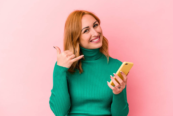 Young caucasian woman holding a mobile phone isolated on pink background showing a mobile phone call gesture with fingers. - 写真・画像