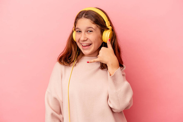 Kleines kaukasisches Mädchen, das isoliert Musik auf rosa Hintergrund hört und eine Handygeste mit den Fingern zeigt. - Foto, Bild
