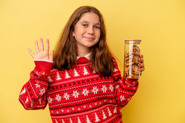 Little caucasian girl holding a Christmas cookies isolated on yellow background smiling cheerful showing number five with fingers. - Fotó, kép