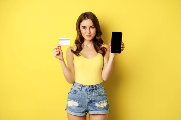 Young beautiful woman model showing smartphone empty phone screen and credit card, ready for summer, wearing tank top and shorts, standing over yellow background - Foto, Bild