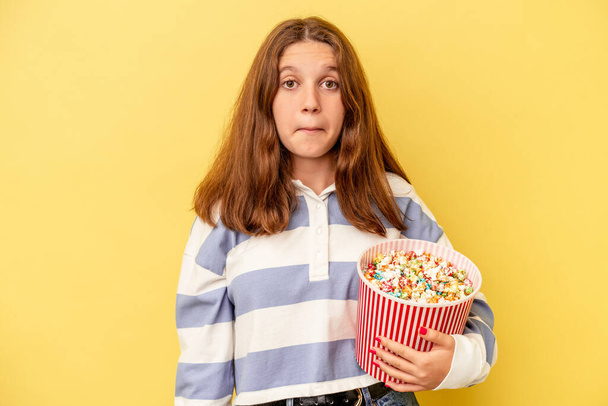 Pieni valkoihoinen tyttö tilalla popcornia eristetty keltainen tausta kohauttaa olkapäät ja avoimet silmät sekava. - Valokuva, kuva