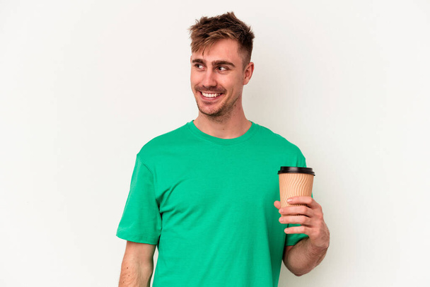 Fiatal kaukázusi férfi kezében egy elvitelre kávé elszigetelt fehér háttér néz félre mosolygós, vidám és kellemes. - Fotó, kép
