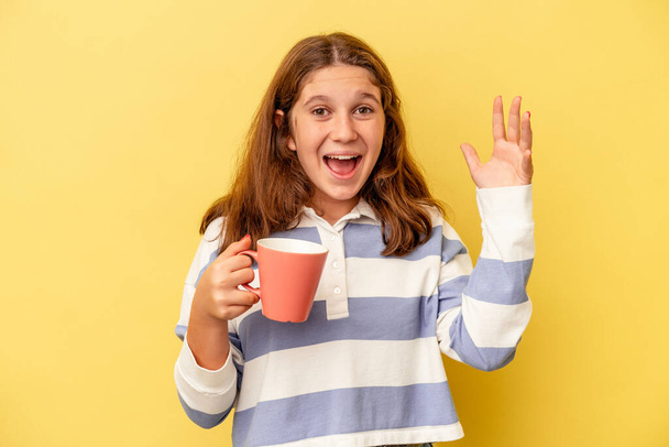 Petite fille caucasienne tenant une tasse rose isolée sur fond jaune recevant une agréable surprise, excitée et levant les mains. - Photo, image