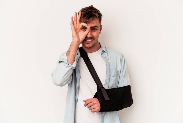 Young caucasian man with broken hand isolated on white background excited keeping ok gesture on eye. - Photo, Image