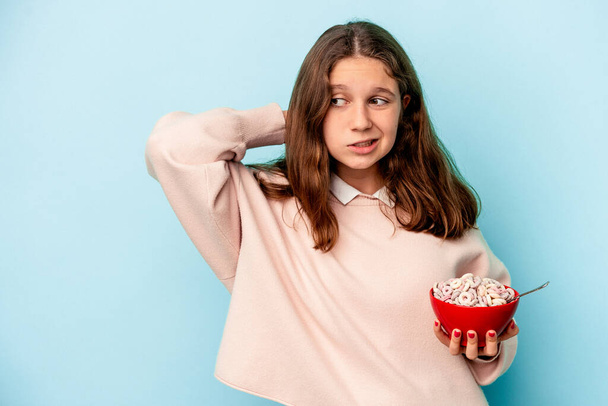 Little caucasian girl holding a bowl of cereals isolated on blue background touching back of head, thinking and making a choice. - 写真・画像