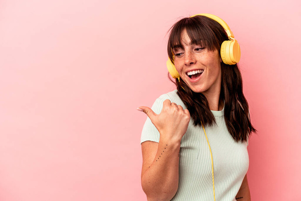 Junge Argentinierin hört Musik isoliert auf rosa Hintergrund zeigt mit dem Daumen weg, lacht und unbeschwert. - Foto, Bild