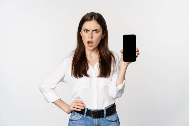 Geschokte en gefrustreerde vrouw tonen mobiele telefoon scherm app, smartphone applicatie, kijken beledigd, staande over witte achtergrond - Foto, afbeelding