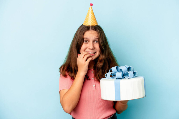 Μικρή καυκάσιος κορίτσι γιορτάζει τα γενέθλιά της κρατώντας μια τούρτα απομονωμένη σε μπλε φόντο τρώει νύχια, νευρικό και πολύ ανήσυχος. - Φωτογραφία, εικόνα