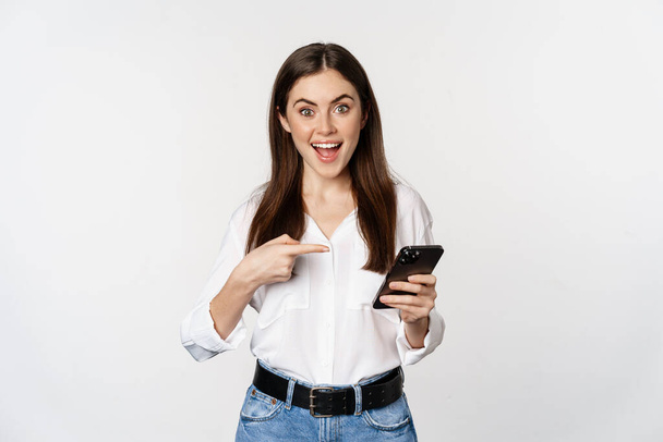 Enthusiastic girl pointing finger at smartphone and smiling, showing big sale online, using app on mobile phone, white background - Φωτογραφία, εικόνα
