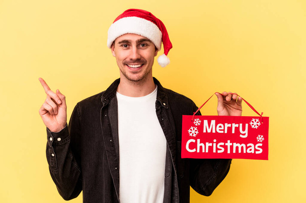 Sarı arka planda izole edilmiş mutlu bir Noel tabelası tutan genç beyaz adam gülümsüyor ve boş alanda bir şeyler gösteriyor.. - Fotoğraf, Görsel