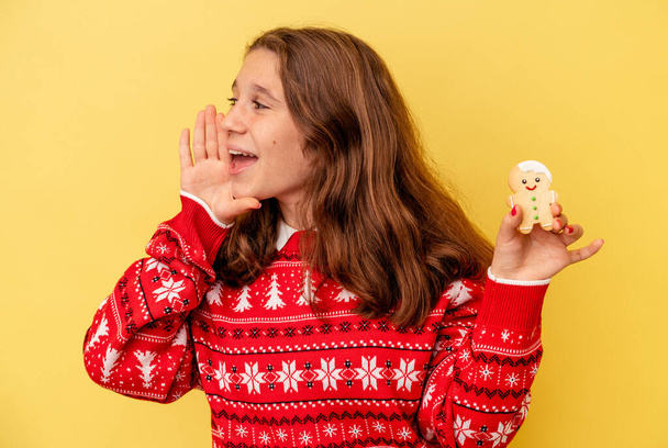Kleines kaukasisches Mädchen mit einem Weihnachtsgebäck auf gelbem Hintergrund, das schreit und die Handfläche in der Nähe des geöffneten Mundes hält. - Foto, Bild