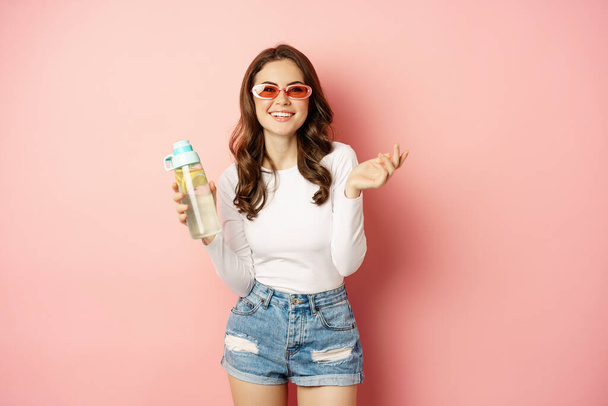 Elegante ragazza in abito primaverile, indossando occhiali da sole, tenendo bottiglia d'acqua con limone, bevanda sana, ridere e sorridere, in piedi su sfondo rosa - Foto, immagini