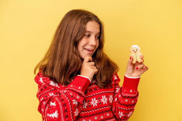 Маленька біла дівчинка тримає різдвяне печиво ізольовано на жовтому тлі, дивлячись в сторону з сумнівним і скептичним виразом
. - Фото, зображення