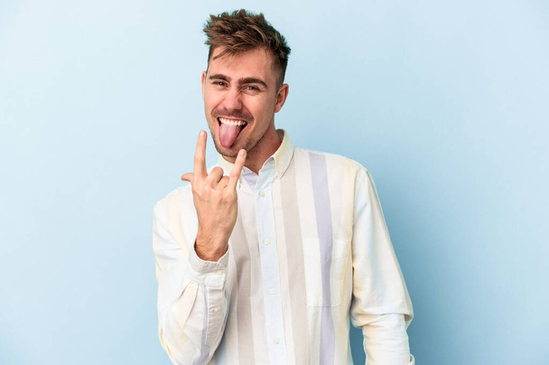 Young caucasian man isolated on blue background showing rock gesture with fingers - Photo, Image