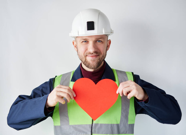Ημέρα του Αγίου Βαλεντίνου: αρσενικό εργαζόμενος σε σκληρό καπέλο δείχνει κόκκινη καρδιά - Φωτογραφία, εικόνα