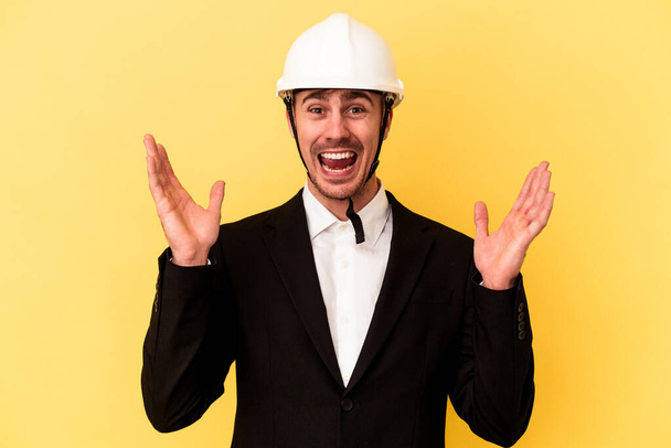 Young architect caucasian man isolated on yellow background receiving a pleasant surprise, excited and raising hands. - Foto, Imagen