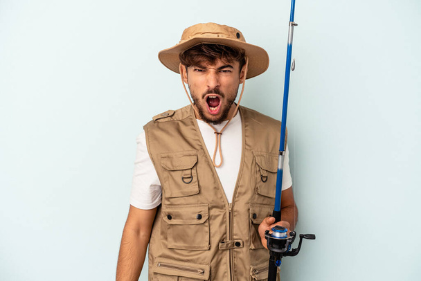 Young mixed race fisherman holding a rod isolated on blue background screaming very angry and aggressive. - Foto, Imagen