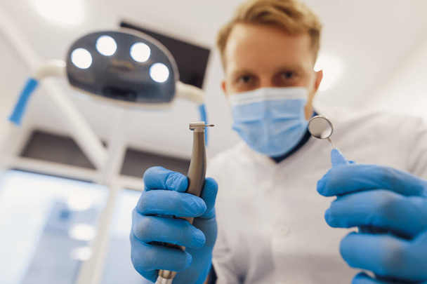 Vista de ángulo bajo del dentista masculino en máscara médica que sostiene las herramientas del dentista para el procedimiento dental en la clínica - Foto, imagen