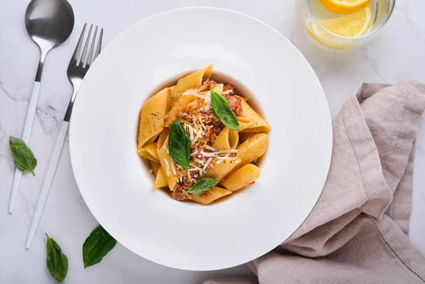 Bolognese Penne Pasta. Tasty appetizing classic italian penne pasta with parmesan cheese, basil and Bolognese sauce in white plate on plate on stone table Traditional dish of Italian cuisine. Top view - Zdjęcie, obraz