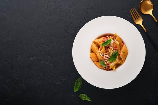 Bolognese Penne Pasta. Tasty appetizing classic italian penne pasta with parmesan, basil and Bolognese sauce in white plate on plate on black dark background.Traditional Italian cuisine. Top view. - Photo, image