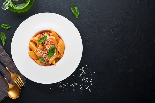 Bolognese Penne Pasta. Tasty appetizing classic italian penne pasta with parmesan, basil and Bolognese sauce in white plate on plate on black dark background.Traditional Italian cuisine. Top view. - Zdjęcie, obraz