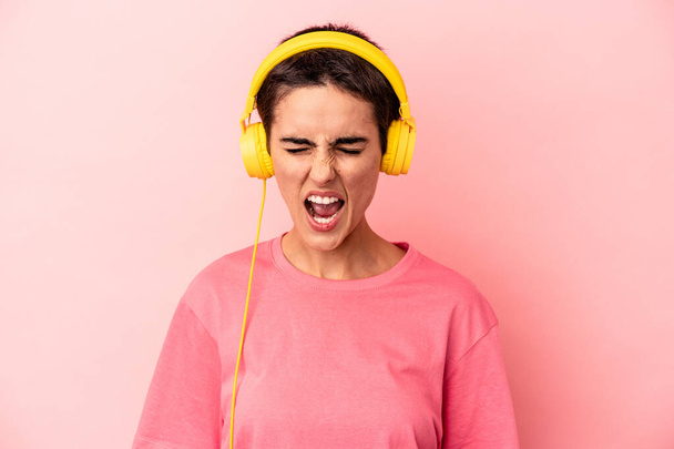 Junge kaukasische Frau hört Musik isoliert auf rosa Hintergrund schreit sehr wütend und aggressiv. - Foto, Bild