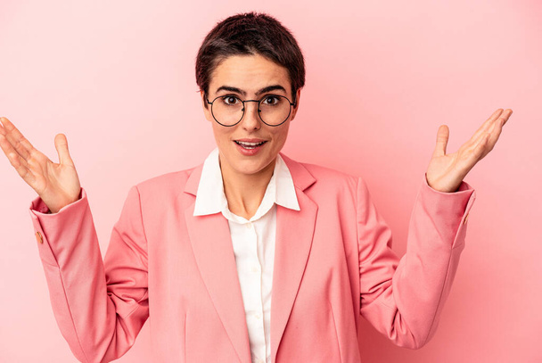 Joven mujer de negocios con una chaqueta rosa aislada sobre fondo rosa recibiendo una agradable sorpresa, emocionada y levantando las manos. - Foto, imagen