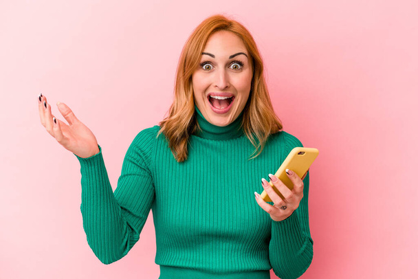 Young caucasian woman holding a mobile phone isolated on pink background receiving a pleasant surprise, excited and raising hands. - Photo, Image