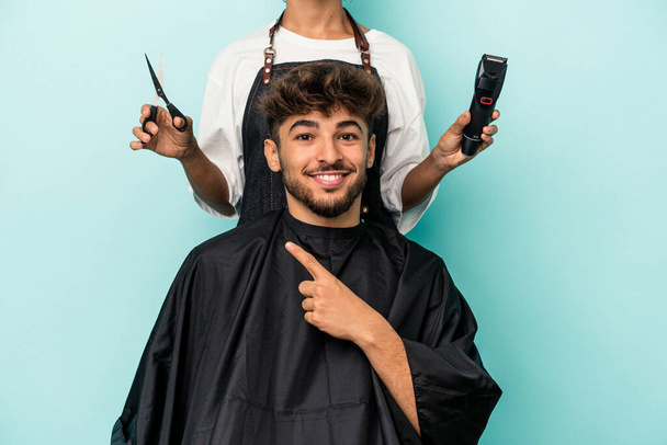 Young arab man ready to get a haircut isolated on blue background smiling and pointing aside, showing something at blank space. - Fotoğraf, Görsel