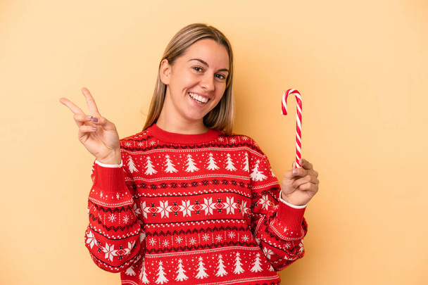 黄色の背景に孤立したクリスマススティックを持つ若い白人女性喜びと屈託のない指で平和のシンボルを示す. - 写真・画像