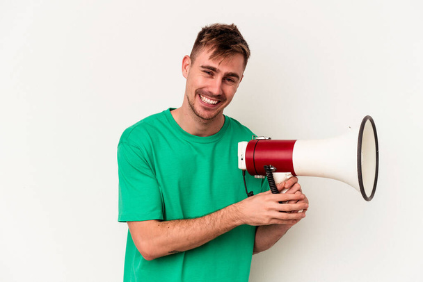 Young caucasian man holding megaphone isolated on white background laughing and having fun. - 写真・画像