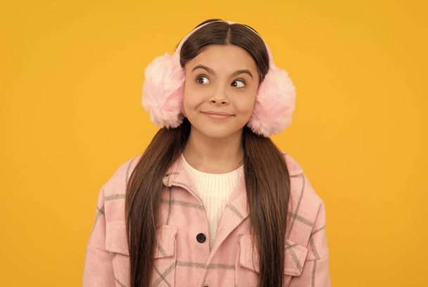 Happy girl child in warm winter fur earmuffs smile with sly look yellow background, intrigue - Foto, imagen