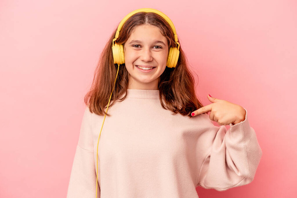 Pequeña chica caucásica escuchando música aislada en el fondo rosa persona señalando a mano a un espacio de copia de la camisa, orgulloso y seguro - Foto, imagen