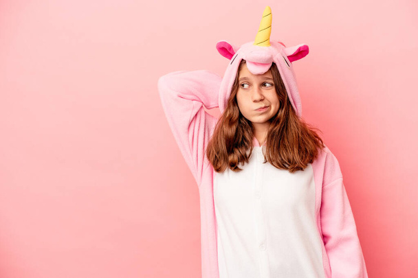 Menina caucasiana vestindo um pijama unicórnio isolado no fundo rosa tocando parte de trás da cabeça, pensando e fazendo uma escolha. - Foto, Imagem